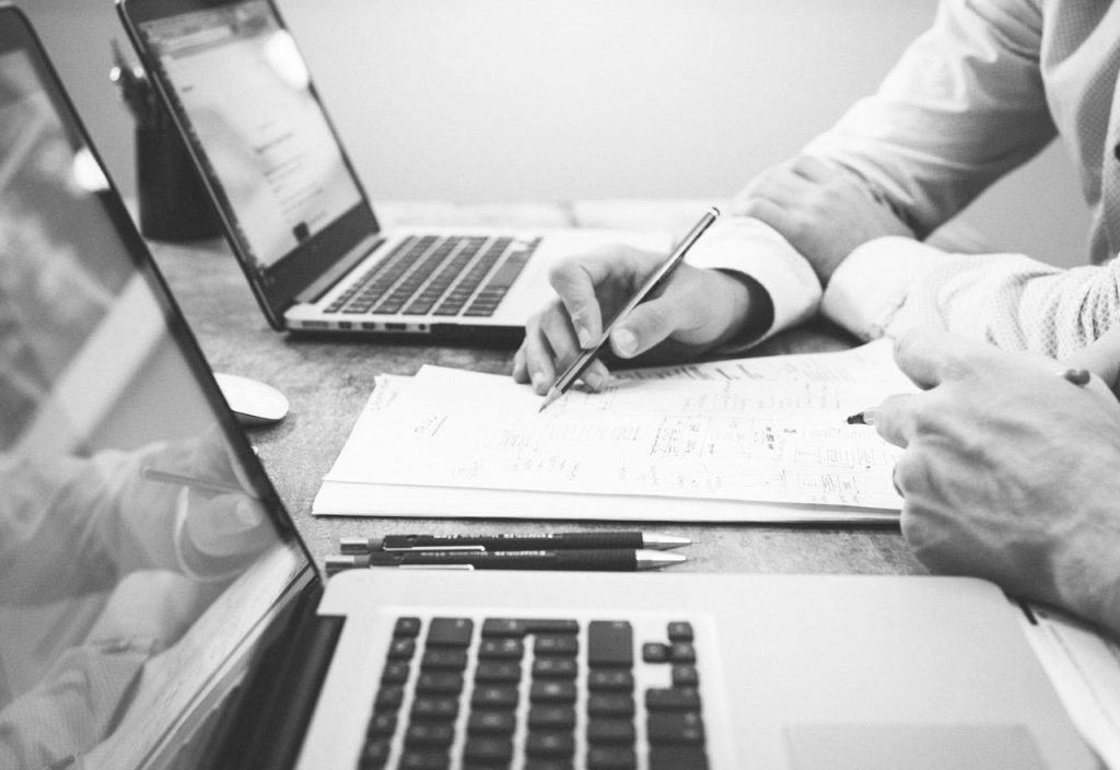 Zwei Personen sitzen vor 2 Laptops und notieren sich handschriftlich Sachen auf einem Zettel in der Mitte